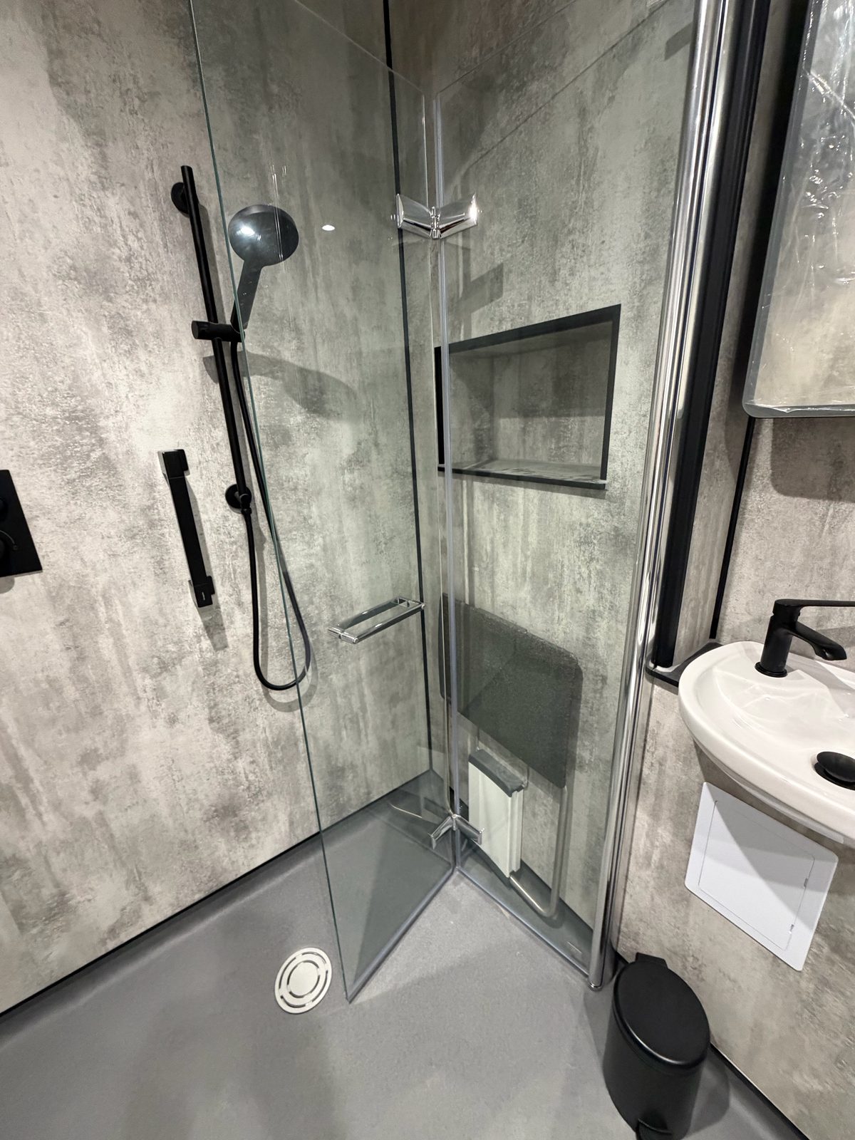 Wet room glass shower open showing fold-down seat and basin against grey wall panels